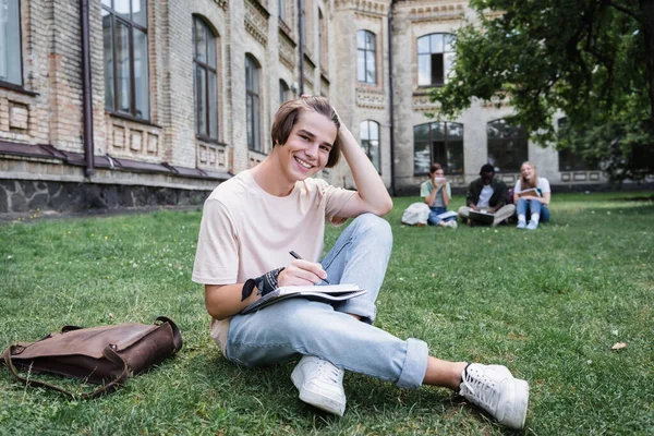 Estudiante alegre escribiendo en el cuaderno y mirando la cámara en el césped - foto de stock