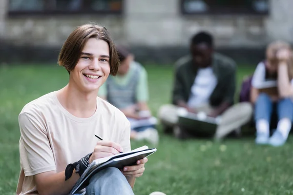 Estudiante feliz mirando la cámara mientras escribe en el cuaderno - foto de stock