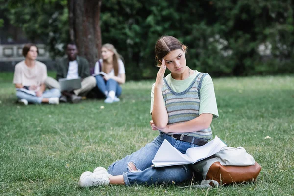 Estudiante molesto con cuaderno y mochila sentado en la hierba en el parque - foto de stock