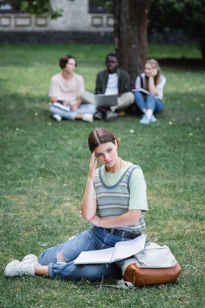 Pensive student with notebook and backpack sitting near blurred friends outdoors — Stock Photo