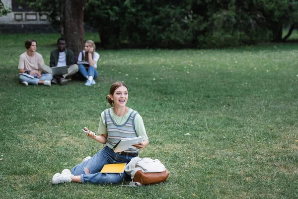 Cheerful student with smartphone and notebook sitting on lawn near blurred interracial friends — Stock Photo