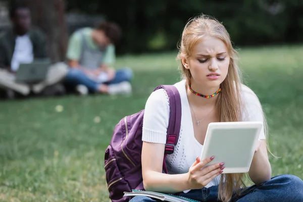 Estudiante molesto con tableta digital y mochila sentada en el césped - foto de stock