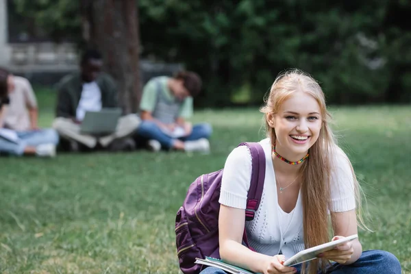 Estudante sorrindo com mochila e tablet digital olhando para a câmera no gramado — Fotografia de Stock