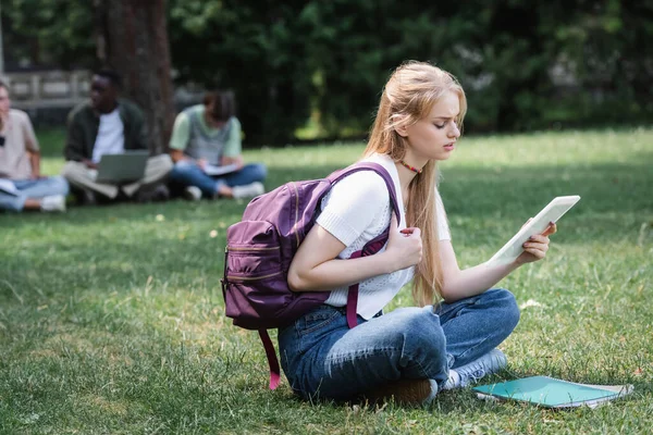 Adolescente estudiante con tableta digital sentado cerca de portátiles en la hierba - foto de stock
