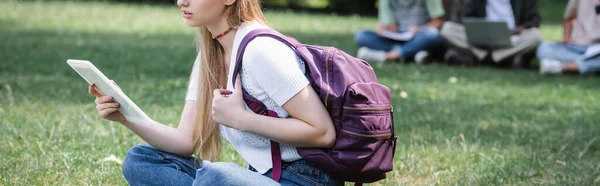 Vista recortada de estudiante sosteniendo la mochila y la tableta digital en la hierba, bandera - foto de stock