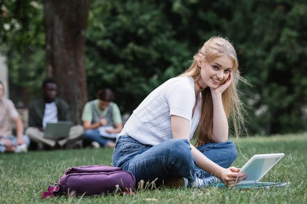 Estudante sorrindo segurando tablet digital e olhando para a câmera no gramado — Fotografia de Stock