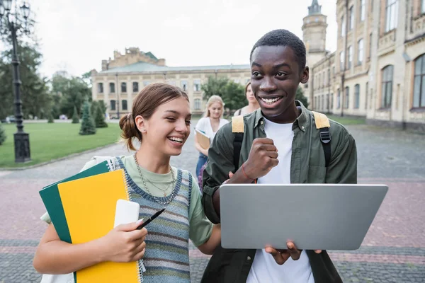 Возбужденный африканский студент-американец показывает да рядом с друзьями с блокнотами на открытом воздухе — стоковое фото