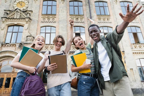 Low angle view of excited interracial students with notebooks looking at camera outdoors — Stock Photo