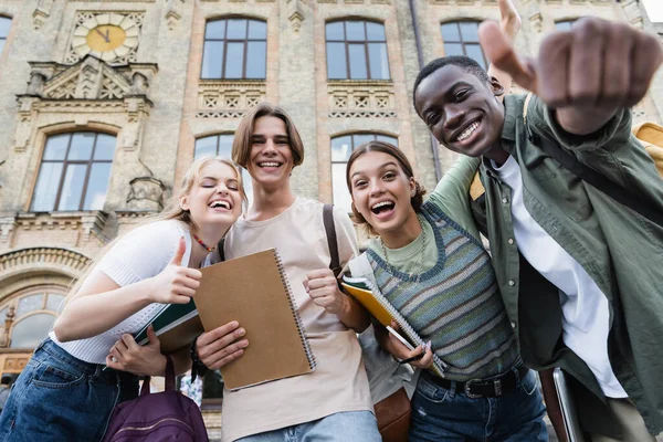 Visão de baixo ângulo de estudantes multiétnicos gesticulando perto de prédio embaçado ao ar livre — Fotografia de Stock