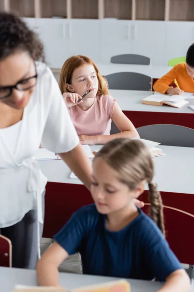 Selective focus of thoughtful redhead girl near blurred multiethnic classmates and african american teacher — Stock Photo