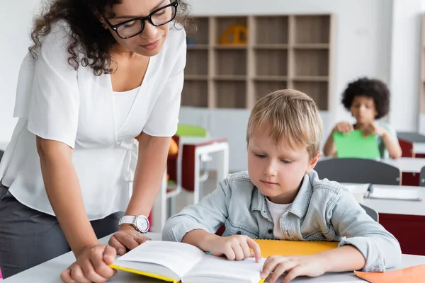African american teacher and schoolboy looking in book during lesson — Stock Photo