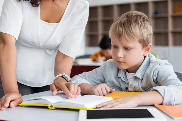 African american teacher pointing with finger at book near schoolboy and digital tablet — Stock Photo