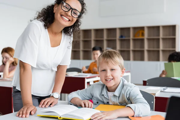 Happy african american teacher and schoolboy looking at camera near book on desk — Stock Photo