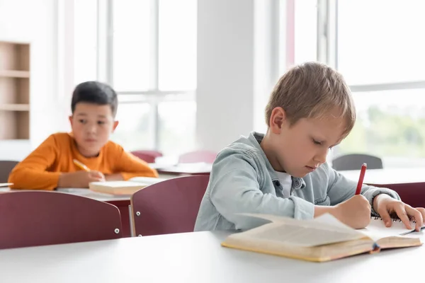 Schoolboy writing during lesson near asian classmate on blurred background — Stock Photo