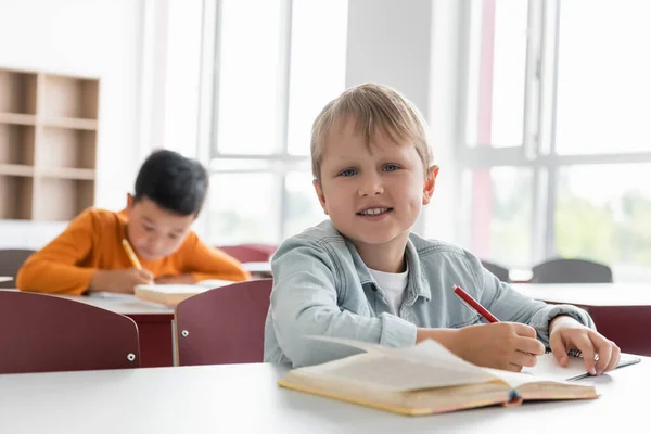 Glückliches Schulkind lächelt in die Kamera neben asiatischem Jungen, der auf verschwommenem Hintergrund schreibt — Stockfoto