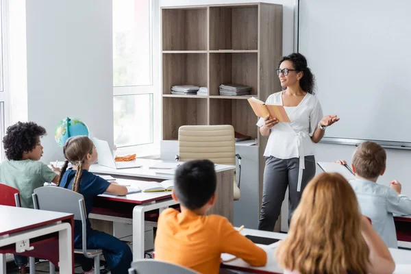 Profesor afroamericano señalando con la mano mientras lee libro en el aula - foto de stock