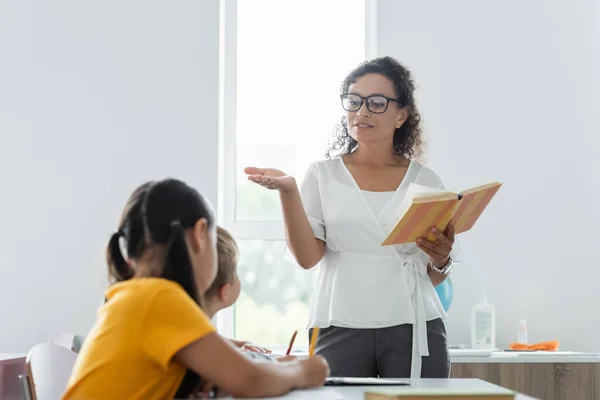 Afrcan professeur américain pointant avec la main tout en tenant le livre près des élèves dans la classe — Photo de stock