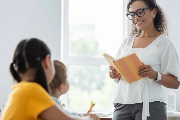 Happy african american teacher reading book to pupils during lesson — Stock Photo