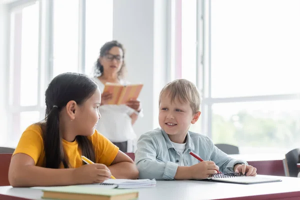 Interracial pupils looking at each other while writing dictation near blurred african american teacher — Stock Photo