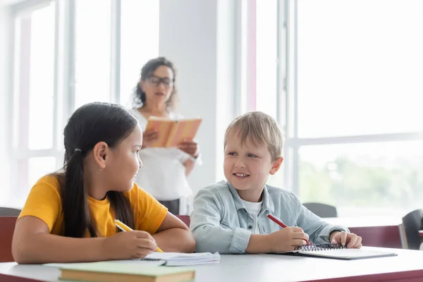 Interrassische Schüler, die einander beim Schreiben von Diktaten fast verschwommen anschauen — Stockfoto