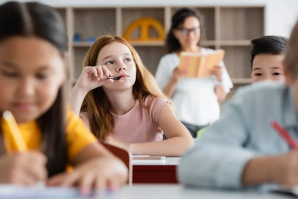 Menina ruiva pensativa segurando caneta na boca enquanto olha para longe durante a lição — Fotografia de Stock