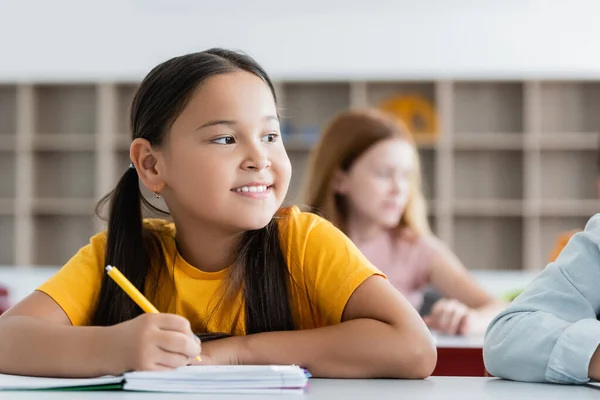 Happy asian girl looking away while holding pen near notebook — Stock Photo