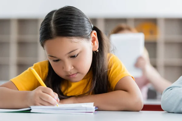 Focused asian schoolgirl writing in notebook during lesson — Stock Photo