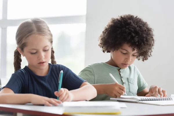 Niños interraciales escribiendo en cuadernos durante la lección - foto de stock