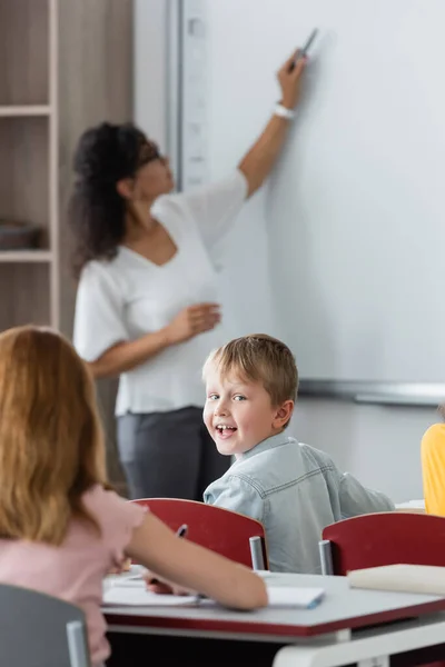 Selektiver Fokus eines glücklichen Jungen, der in die Kamera schaut, in der Nähe verschwommener afrikanisch-amerikanischer Lehrer, der auf Whiteboard schreibt — Stockfoto