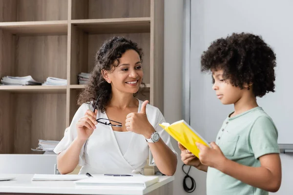 Niño afroamericano leyendo libro a profesor sonriente durante la lección - foto de stock