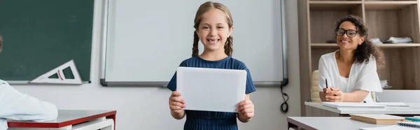 Щаслива школярка тримає цифровий планшет біля вчителя, посміхаючись у класі на розмитому фоні, банер — Stock Photo