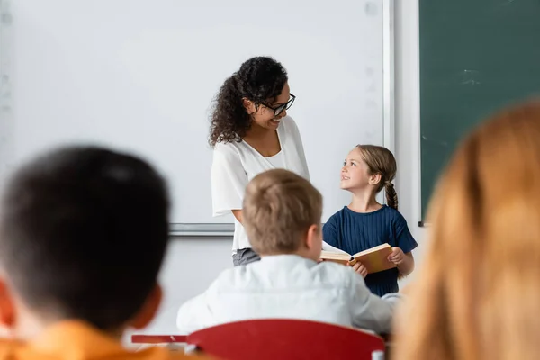Menina com livro e professor afro-americano positivo olhando um para o outro enquanto em pé na sala de aula — Fotografia de Stock