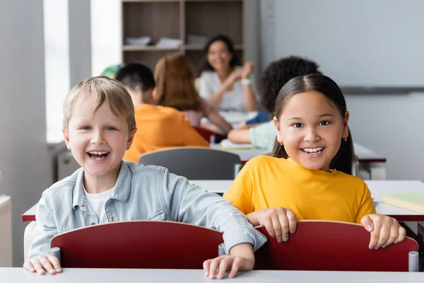 Cheerful interracial schoolkids looking at camera near blurred classmates and african american teacher — Stock Photo