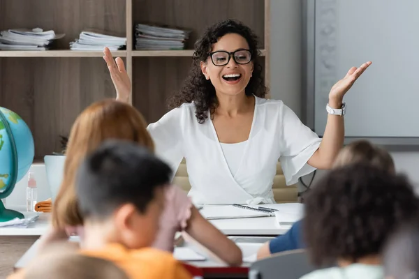 Excited african american teacher showing wow gesture near blurred children in classroom — Stock Photo