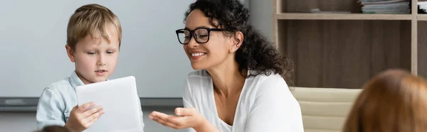 Cheerful african american teacher pointing at digital tablet near schoolboy, banner — Stock Photo