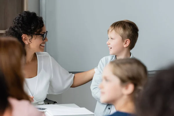 Positivo africano americano insegnante parlando con scolaro durante lezione vicino sfocato alunni — Foto stock