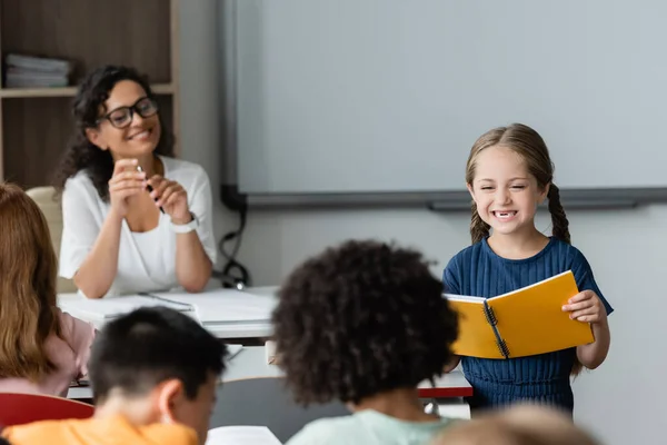 Joyeuse écolière tenant un cahier tout en étant debout dans la salle de classe pendant la leçon — Photo de stock