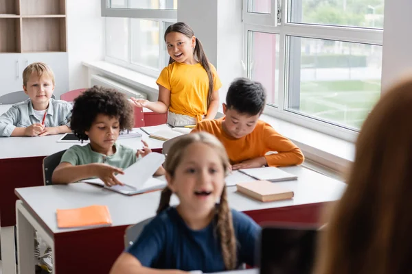 Positivo asiático menina gestos enquanto falando perto multiétnico alunos em sala de aula — Fotografia de Stock