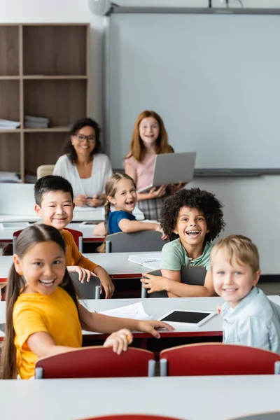 Alegre multicultural niños mirando a la cámara mientras africano profesor sonriendo en borrosa fondo - foto de stock