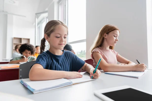 Niños multiculturales escribiendo en cuadernos durante la lección - foto de stock