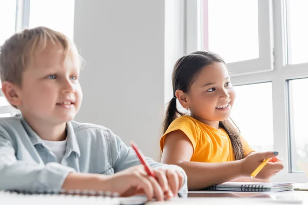 Happy interracial schoolkids holding pens during lesson in classroom — Stock Photo