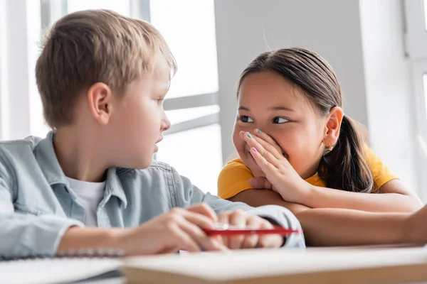 Asian girl covering mouth with hand while telling secret to classmate — Stock Photo