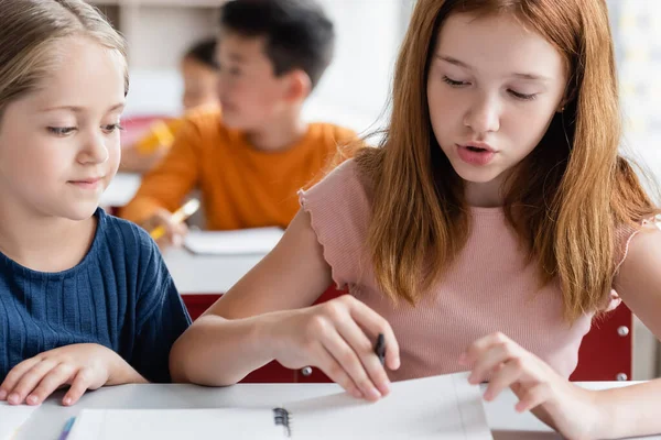 Colegialas cerca de cuaderno y difuminado compañeros de clase multiétnicos en el aula - foto de stock