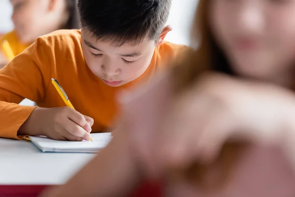 Asiático chico escritura en notebook cerca borrosa compañeros de clase - foto de stock