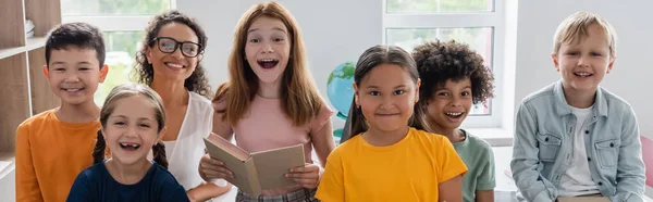 Chica emocionada sosteniendo libro cerca de compañeros de clase multiétnicos felices y profesor afroamericano, pancarta - foto de stock