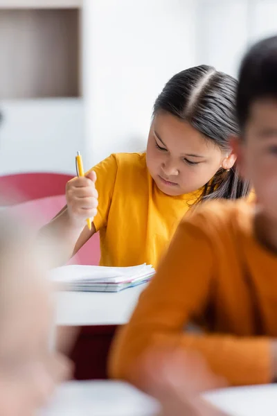 Asiático chica celebración pluma cerca notebook y borrosa compañeros de clase - foto de stock