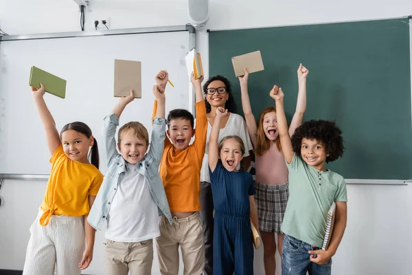 Estudiantes multiétnicos emocionados sosteniendo libros en manos levantadas cerca del feliz profesor afroamericano - foto de stock