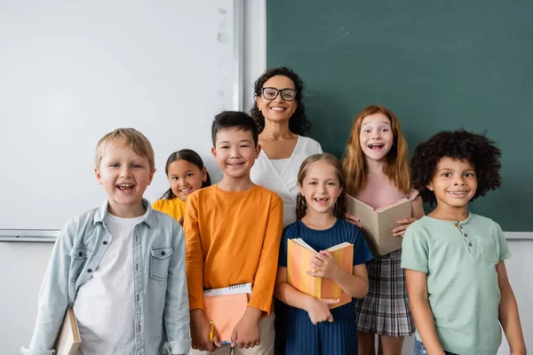 Enseignant afro-américain et joyeux écoliers multiethniques souriant à la caméra dans la salle de classe — Photo de stock