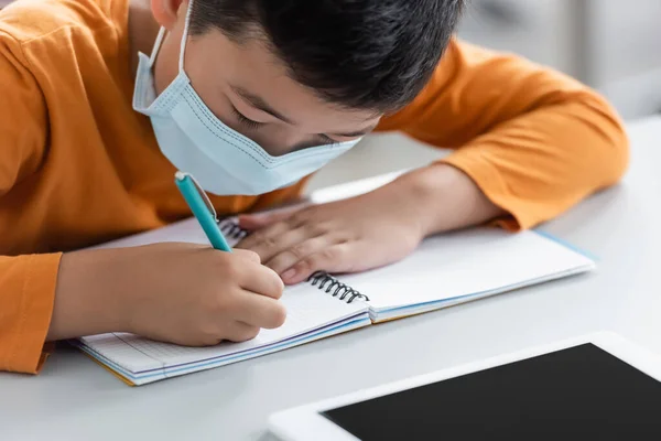 Азиатский мальчик в медицинской маске записи в ноутбуке рядом с цифровой планшет — стоковое фото