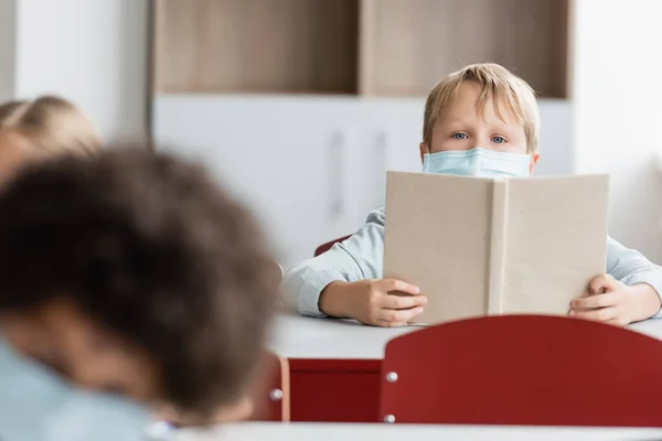 Estudante em máscara médica segurando livro enquanto olha para a câmera durante a aula — Fotografia de Stock